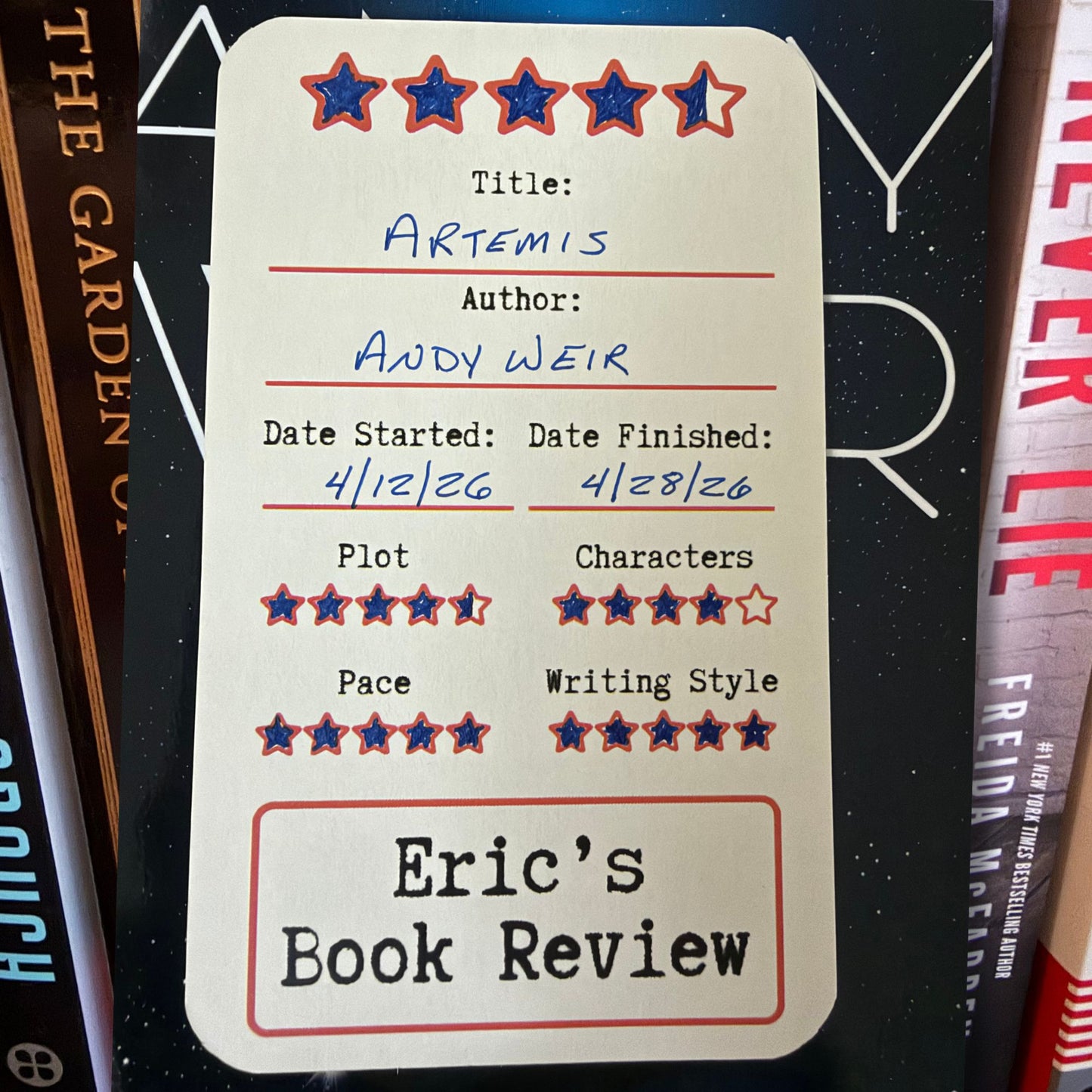 A cream-colored book review card with red and blue star ratings and handwritten text for title, author, start and finish dates, plus quality scores, photographed standing upright among book spines.