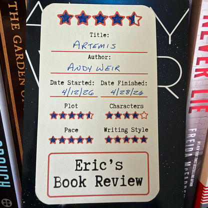 A cream-colored book review card with red and blue star ratings and handwritten text for title, author, start and finish dates, plus quality scores, photographed standing upright among book spines.